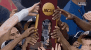 Sports gif. The UConn men's basketball team celebrate their win at the NCAA championship, crowding together as they smile and hold up their trophy.