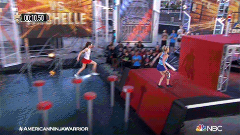 Reality TV gif. Two women race each other up a wall on American Ninja Warrior. The girl in the blue shirt pushes the button first and the girl in the white shirt collapses. The blue shirt woman collapses on top of the other girl.