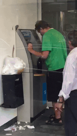 Video gif. Man stands unsteadily at an ATM with a bottle on the machine. He accidentally knocks the bottle off and it falls landing straight up, spraying the man right in the face with foamy alcohol. People around him jump back.
