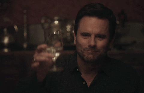TV gif. Deacon Claybourne as Charles Esten sits at a dining table holding a glass of water in his hand. He looks down the table with a warm smile, and lifts his glass in celebration. He says, “Cheers.”