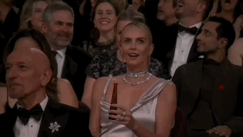 Oscars 2024 GIF. Charlize Theron, seated at the Oscars, looks about, laughing in surprise, saying, “Wow!”