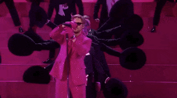 Oscars 2024 GIF. Ryan Gosling is in the middle of his performance for I'm Just Ken and he puts the microphone back on the stand. The microphone stand is way too tall and Gosling shortens it, bringing it down to his height. 