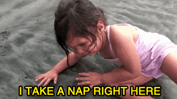 Video gif. A little girl in a swimsuit lies down on wet sand. Text, "I take a nap right here."