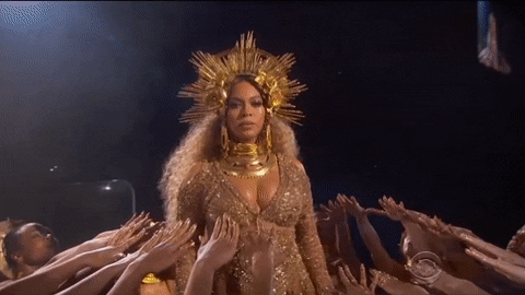 Celebrity gif. Dressed in a gold outfit and headpiece, Beyonce stands looking stoic as she is surrounded by many women who are holding up their hands to her in worship.
