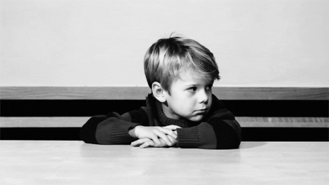 Video gif. A little boy sits with his arms crossed on a table. He turns his head to the side, crinkling his brow in boredom and tapping his fingers impatiently on his arm.