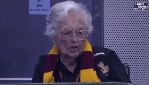 Sports gif. Old woman attentively watches basketball game from audience while taking bites of popcorn.