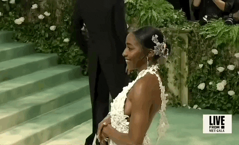 Met Gala 2024 gif. Ayo Edibiri wearing an unfussy Loewe gown of white 3D flower cutouts that fade into color, poses for the cameras, swinging one way, then the other, adjusting poses and offering cheeky looks.
