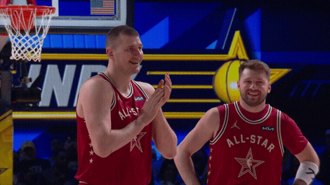 Sports gif. Luka Doncic and Nikola Jokic of the Dallas Mavericks and Denver Nuggets wearing the same red all stars jerseys on the court at the 2024 NBA All-Star Game. Jokic is rubbing his hands together as he smiles and nods at someone offscreen. Doncic has his hands on his hips, smiling and looking amused. 