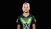 Sports gif. Pernille Harder looks straight at us, then turns to her right, smiles, and points upward with both hands. 