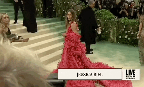 Met Gala 2024 gif. Side angle wide shot of Jessica Biel posing for pictures, wearing a hot pink Tamara Ralph Couture full-length gown with a deep-v neckline. The full billowing skirt flares out to a dramatic train. She wears a billowing shrug that matches the pattern of her dress around her back and arms. 