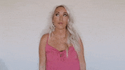 Video gif. Blond woman looks up and smiles, then points up with both of her hands.