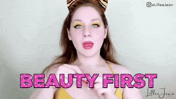 Video gif. Youtuber Lillee Jean looks at us as she pulls a lip gloss wand out of the bottle and applies it to her lips. Her hair is done up and she already has eye shadow and mascara. Text, "Beauty first."