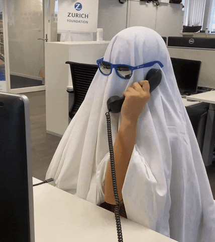 Video gif. In an office a person wears a white sheet over their body and blue sunglasses. They hold up an office phone and nod absentmindedly. 