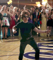Video gif. A man in a black T-shirt and pants swinging his hips on a dance floor in a high school gym, surrounded by students in formal wear.