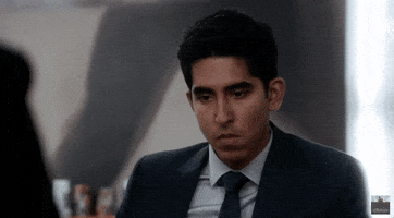 Celebrity gif. Dev Patel eyes cast down and disengaged, bites his lip, considering.