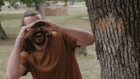TV gif. Jonathan Van Ness from Queer Eye steps out from behind a tree, spying at something in the distance with binoculars. Then he smiles and shakes his head, stepping back behind the tree.