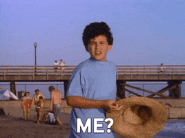 Video gif. A young boy points to himself, confused, as he stops on the beach next to a boardwalk. "Me?" he asks, which appears as text.