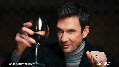 TV gif. Dylan McDermott as Richard, Tamara Taylor
 as Angela and Robin Taylor as Sebastian in Law & Order: Organized Crime. They all sit at a table and smile and toast one another for a job well done.
