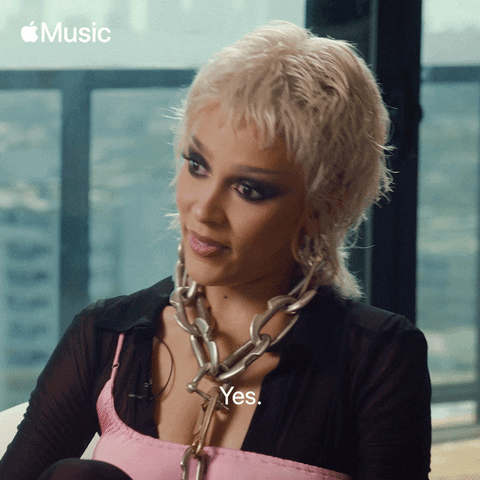 Celebrity gif. Doja Cat says yes, and then her eyes dart around, looking at us and then left and right as she smiles slightly.