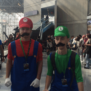 Video gif. People cosplaying as Mario and Luigi at Comic Con hop like their characters. 
