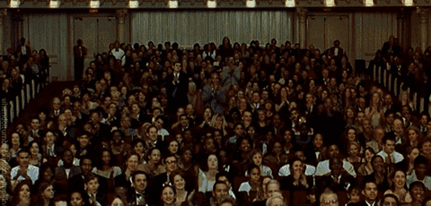 Video gif. In an ornate performance hall a well dressed crowd stands with a standing ovation. 
