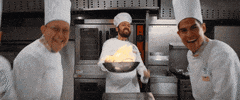 Movie gif. In a restaurant kitchen, men in white chefs' outfits smile at us, standing before a central chef holding a flaming wok and raising his other arm triumphantly.