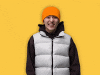 Celebrity gif. Kino from KPOP group Pentagon wears an orange beanie and white vest. He puts both his hands up to his mouth as he giggles mischeviously. 