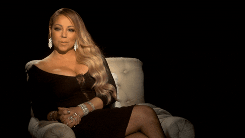 Celebrity gif. Mariah Carey is sitting in a sofa and sends us an air kiss. A golden digital lip print appears in the corner as she kisses the air, sparkling next to her.