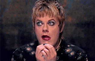 Documentary gif. Eddie Izzard in Dress to Kill wears dark lipstick and eyeliner that accentuates her blue eyes. Her hands are squeezed together as she glances around shiftily and appears doubtful.