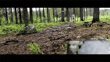 Video gif. Very fit man runs down a trail in a wooded area.