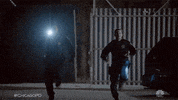 TV gif. Two officers from Chicago PD are running in the dark with their flashlights brandished.