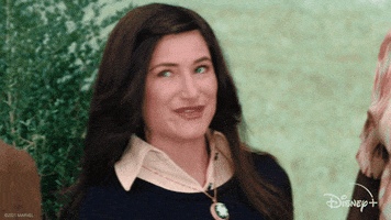 TV gif. From WandaVision: Agnes Harkness (Kathryn Hahn) gives us an exaggerated wink and tilts her head with a smile.