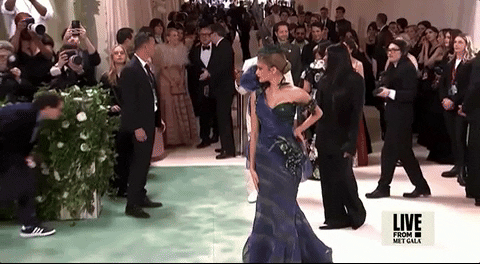 Met Gala 2024 gif. Zendaya wearing a peacock-colored Maison Margiela gown by John Galliano embellished with true-to-life grapes and hummingbirds, struts down the carpet striking pose after pose, sticking the landing with brooding poise.