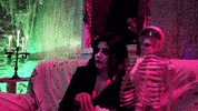 Video gif. Woman wearing a vampire costume eats popcorn on a couch next to a skeleton wearing sunglasses as a creepy monster emerges from the window behind them and waves at us.