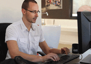 Video gif. A man typing and focusing on his computer and appearing to have three arms, as he takes a sip of his coffee without stopping.