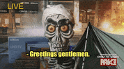 Video gif. Jeff Dunham's Achmed the Dead Journalist, a skeleton puppet, reports from a burning city, a tent and a UHD TV box behind him, raising his eyebrows as he talks to us, saying, "Greetings gentlemen."  