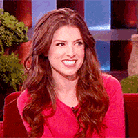 Celebrity gif. Anna Kendrick smiles wide and waves as if she is excited to see someone.