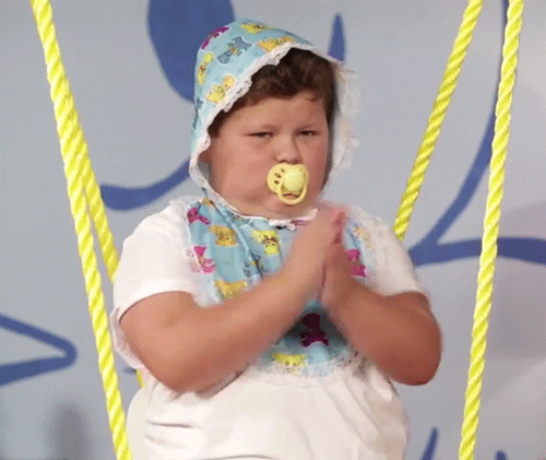 Video gif. Boy dressed as a baby with a bib, a bonnet and a pacifier claps steadily as he sits in a swing.