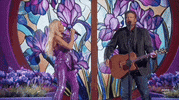 Academy of Country Music Awards gif. Gwen Stefani and Blake Shelton perform on the purple, flower decorated stage as a couple. Blake strums the guitar strapped around his body and sings into the standing mic as he turns his head to lovingly look at Stefani. She matches his gaze while singing into a handheld microphone.