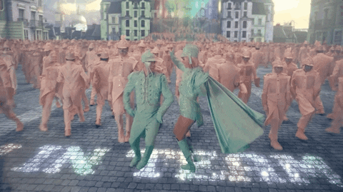 Music video gif. In the video for Me, Taylor Swift circles around Brendan Urie wearing mint-colored suits at the front of a cobblestone street full of dancers wearing pale salmon-pink suits.