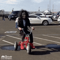 Celebrity gif. Dressed as Jigsaw from the Saw movies, Steph Curry rides a red tricycle slowly through a parking lot, glancing over his shoulder.