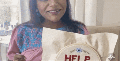 Celebrity gif. Mindy Kaling smiling and showing her embroidery, which reads, "Help, I'm going insane."