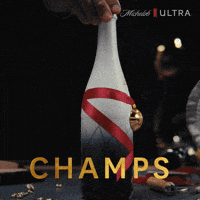 Sponsored gif. Hand turns a fancy champagne bottle of Michelob ULTRA beer so it faces forward. It’s tied with a red ribbon and a gold medal. Glimmering text over it reads, “Champs.”