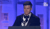 Video gif. Saturday Night Live's Colin Jost stands at a podium at the 2024 White House Correspondents' Dinner and takes a sip of water from a glass as he raises his eyebrows and grimaces, as though to say "the show must go on."