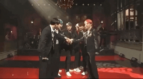 SNL gif. We see BTS standing in a circle with their hands all in. One band member holding a mic flashes a peace sign at us. 