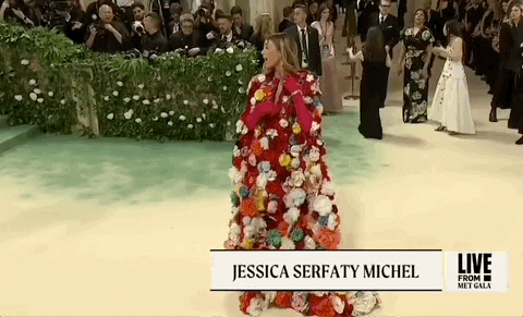 Met Gala 2024 gif. Jessica Serfaty Michel wearing a red Dolce and Gabbana cape covered in large 3D flowers, arrives and adjusts her ensemble to reveal a plunging halter bodice.
