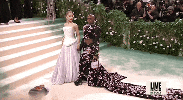 Met Gala 2024 gif. Ariana Grande and Cynthia Erivo turn to look over their shoulders to and away from us pose for a photo. Grande is wearing a pale pink Loewe gown featuring a corset-style bodice with mosaic-like detailing, with draping fabric at the mid section and a flowing pleated skirt of varying colors. Erivo is wearing a two-piece Thom Browne deconstructed tuxedo and skirt featuring a cropped jacket and skirt accented with black sequins and delicate pale pink petal appliqués.