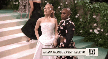 Met Gala 2024 gif. Ariana Grande and Cynthia Erivo pose together to show the front of their outfits. Grande is wearing a pale pink Loewe gown featuring a corset-style bodice with mosaic-like detailing. She is wearing ethereal petal wings on her temples. Erivo is wearing a two-piece Thom Browne deconstructed tuxedo and skirt featuring a cropped jacket and skirt accented with black sequins and delicate pale pink petal appliqués.