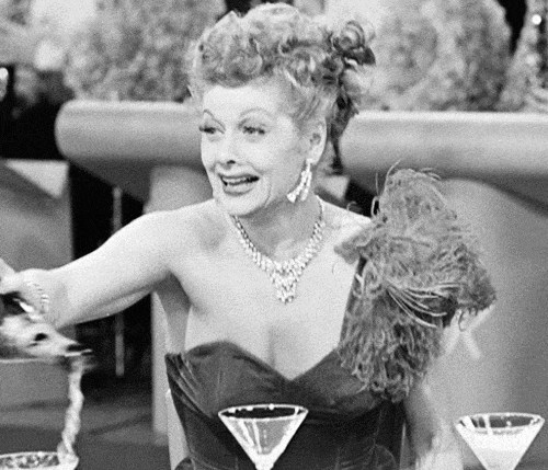 TV-gif.  Lucille Ball van I Love Lucy, gekleed in een mooie jurk en opvallende sieraden, giet opgewonden en lukraak champagne over glazen en de tafel.