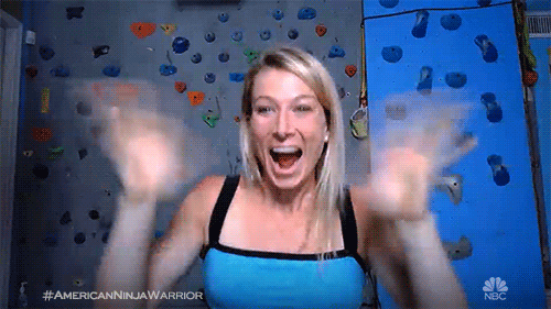 TV gif. Woman from American Ninja Warrior excitedly waves at us with both hands as if in a video call, a rock climbing wall in the background.